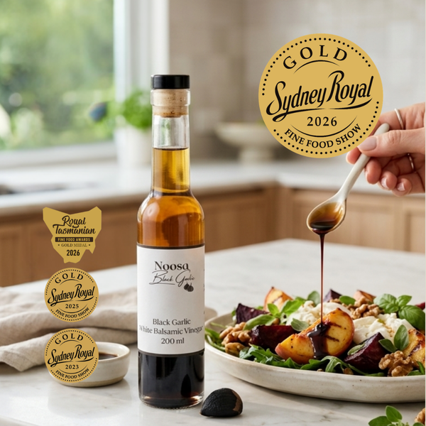 A bottle of Noosa Food Works Black Garlic Balsamic Vinegar stands on a kitchen counter beside a plated salad with roasted vegetables, with a hand holding a spoon to pour the dark balsamic. Three Sydney Royal award badges are shown on the left, and a gold Sydney Royal 2026 badge is shown on the right.
